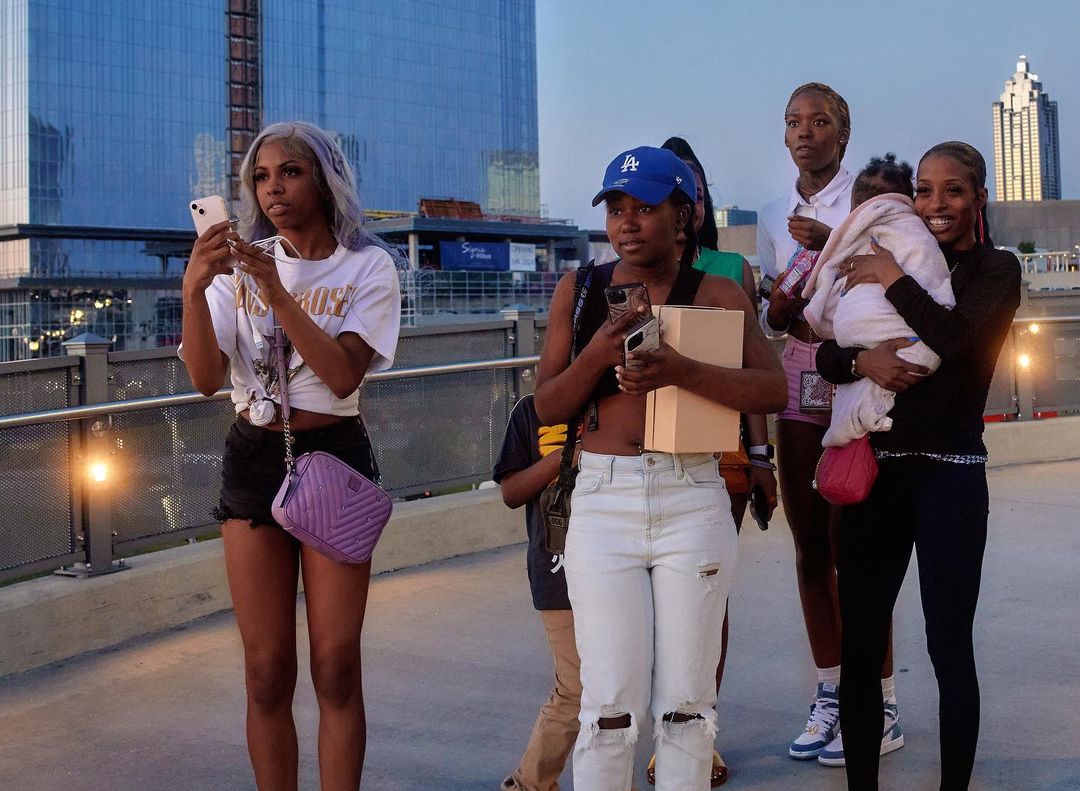 Group of women and children stand on an outdoor walkway in front of modern buildings at dusk, with one woman holding a phone.