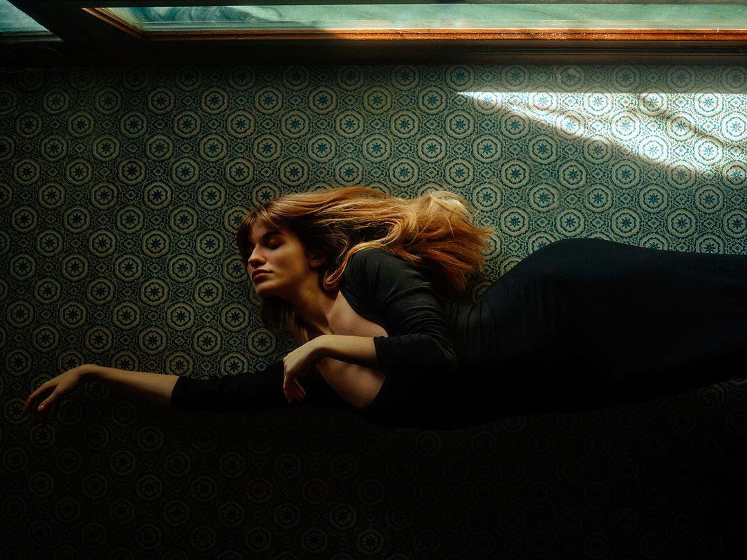 Woman with auburn hair lies on patterned floor in long-sleeve black dress, lit by window light in an indoor setting. 