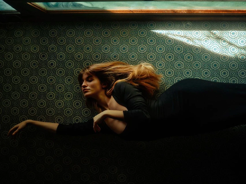 Woman with auburn hair lies on patterned floor in long-sleeve black dress, lit by window light in an indoor setting. 
