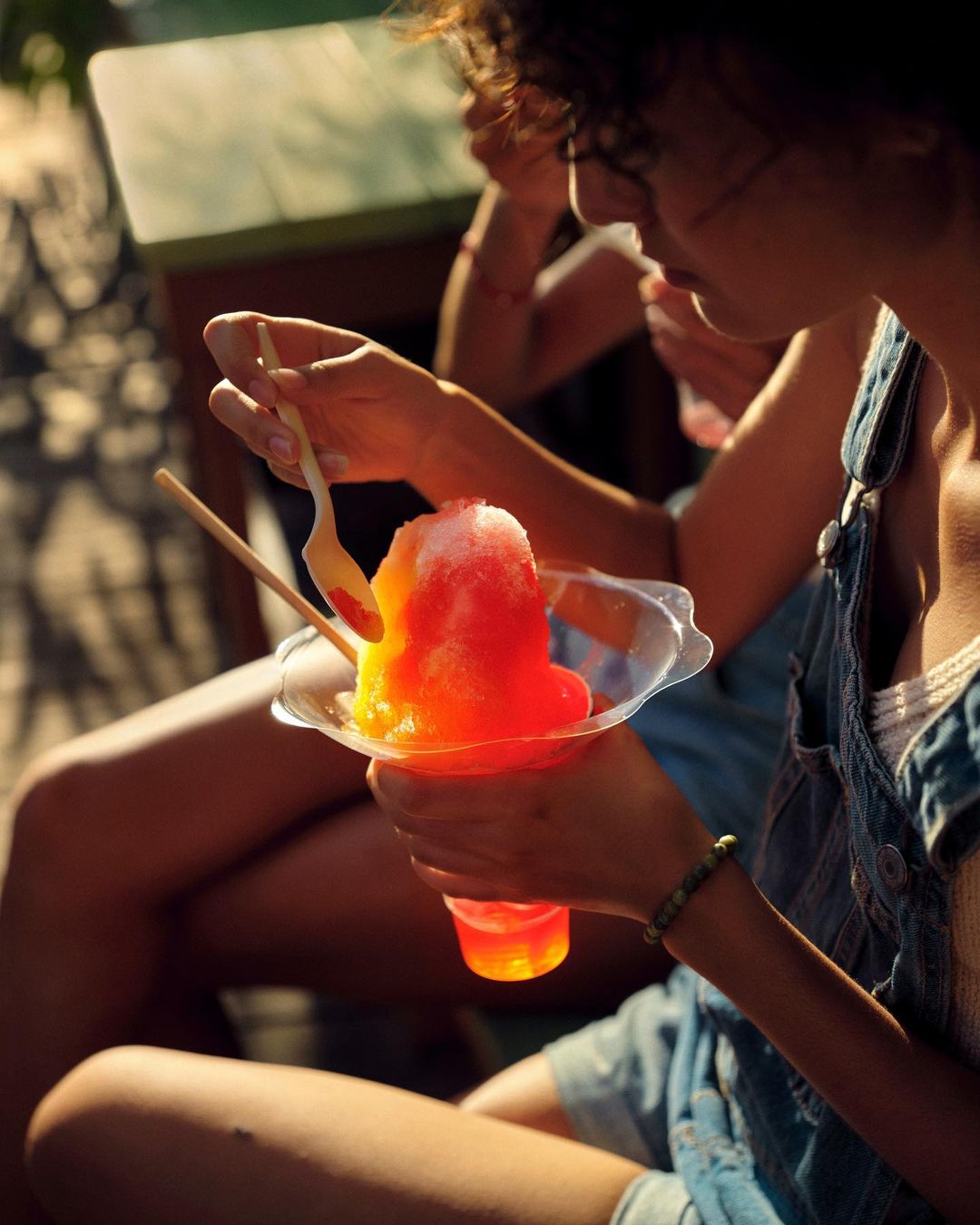 Person enjoying shaved ice outdoors, holding a clear plastic cup filled with red and yellow ice with a small spoon.