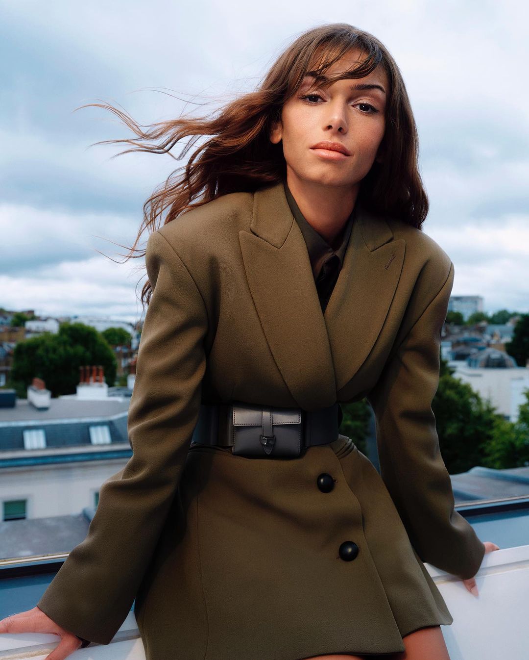Fashion portrait of a woman with flowing hair in a blazer dress, outside on a rooftop with cloudy skies and city buildings.