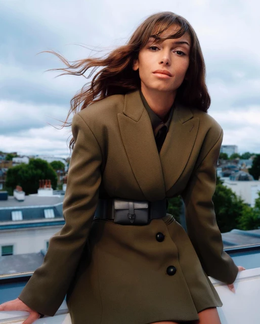 Fashion portrait of a woman with flowing hair in a blazer dress, outside on a rooftop with cloudy skies and city buildings.