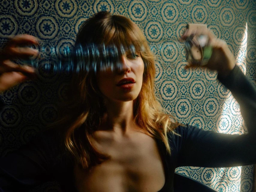 Portrait of a blonde adult woman holding a blurry object in her hands with a patterned wallpaper backdrop in an indoor setting.