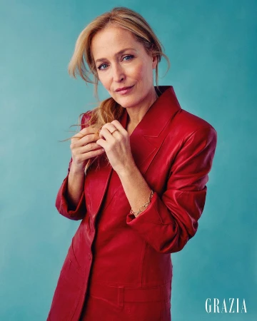 Gillian Anderson wearing a bright red leather blazer against a mottled blue textured studio background.