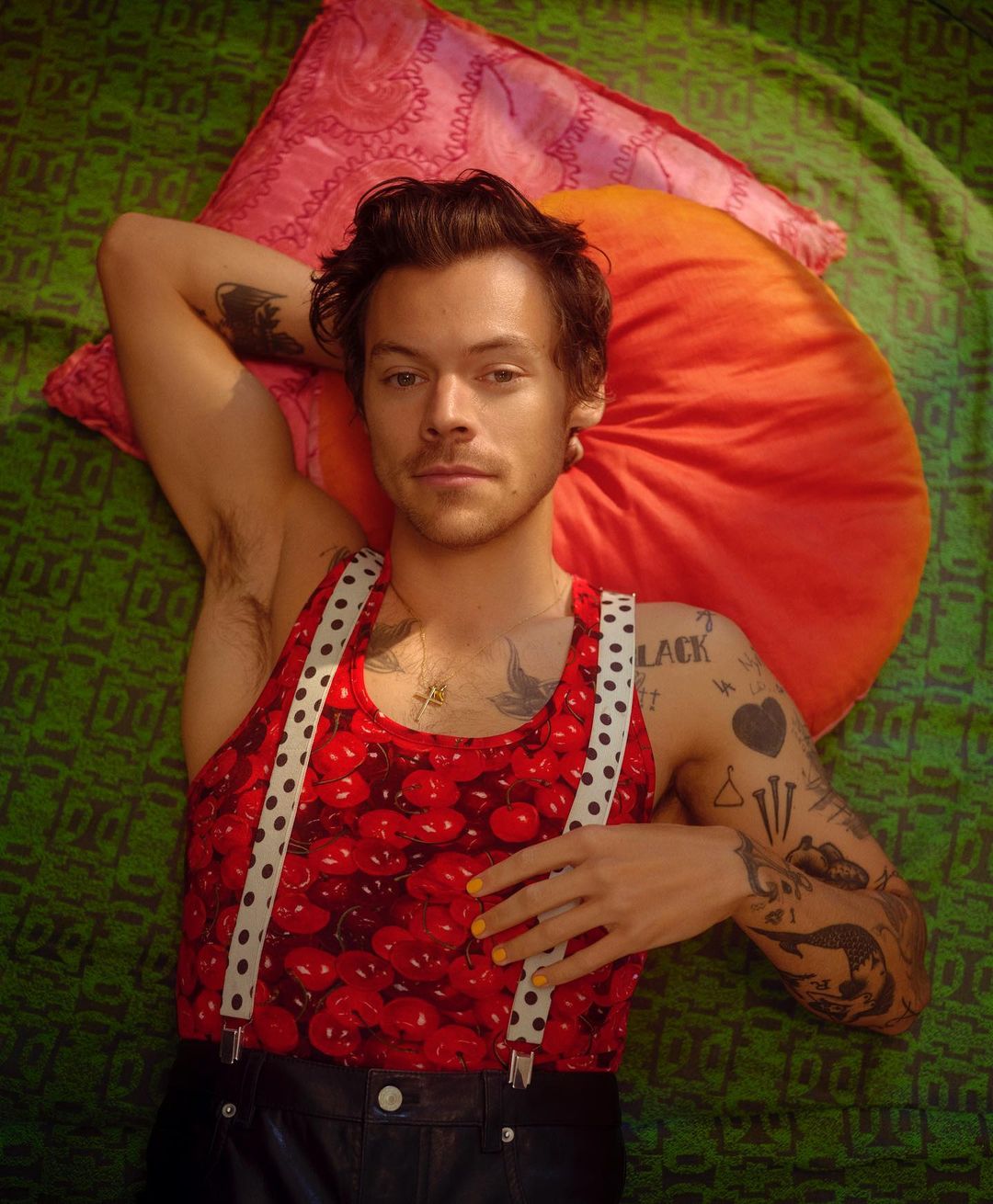 Portrait of a fair-skinned adult male lying down wearing a red tank top and black pants, resting on colorful pillows on a green patterned surface.