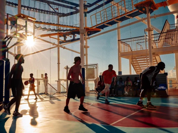 Outdoor basketball court scene with several young adult men playing on a red and blue court, with elevated structures in the background.