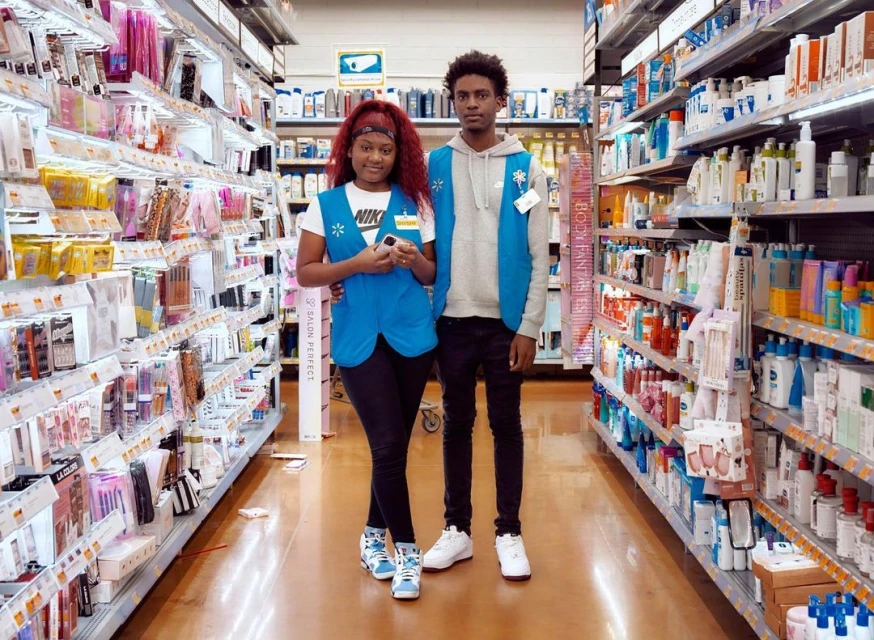Two retail employees in blue vests pose in a brightly lit store aisle surrounded by shelves stocked with toiletries and cosmetics.