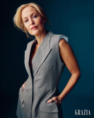 Portrait of Gillian Anderson wearing a sleeveless grey blazer with her hands in the pockets against a dark blue studio background.