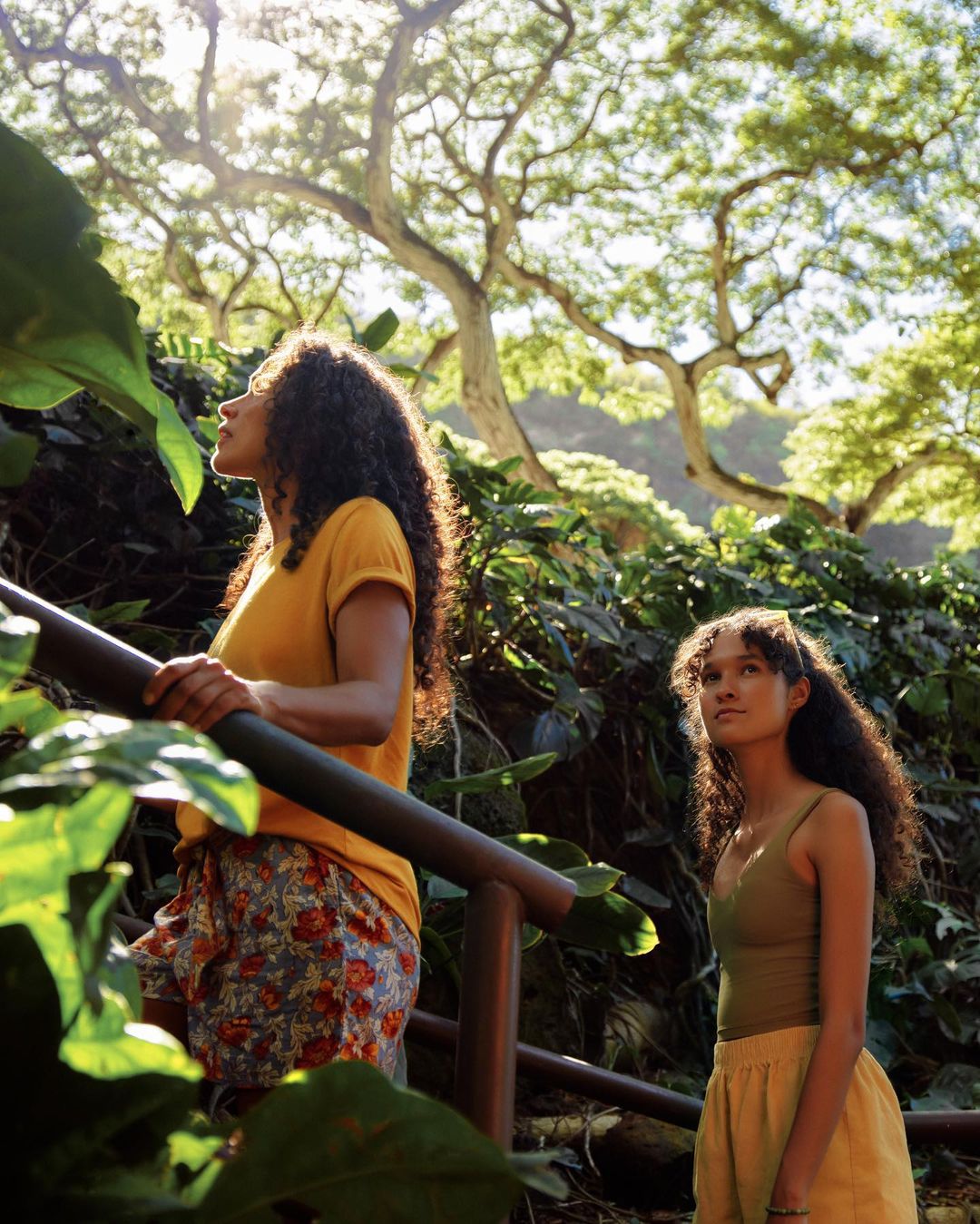 Two young adult women walking up steps in a lush forest setting, bathed in sunlight with the focus on nature and the path ahead.