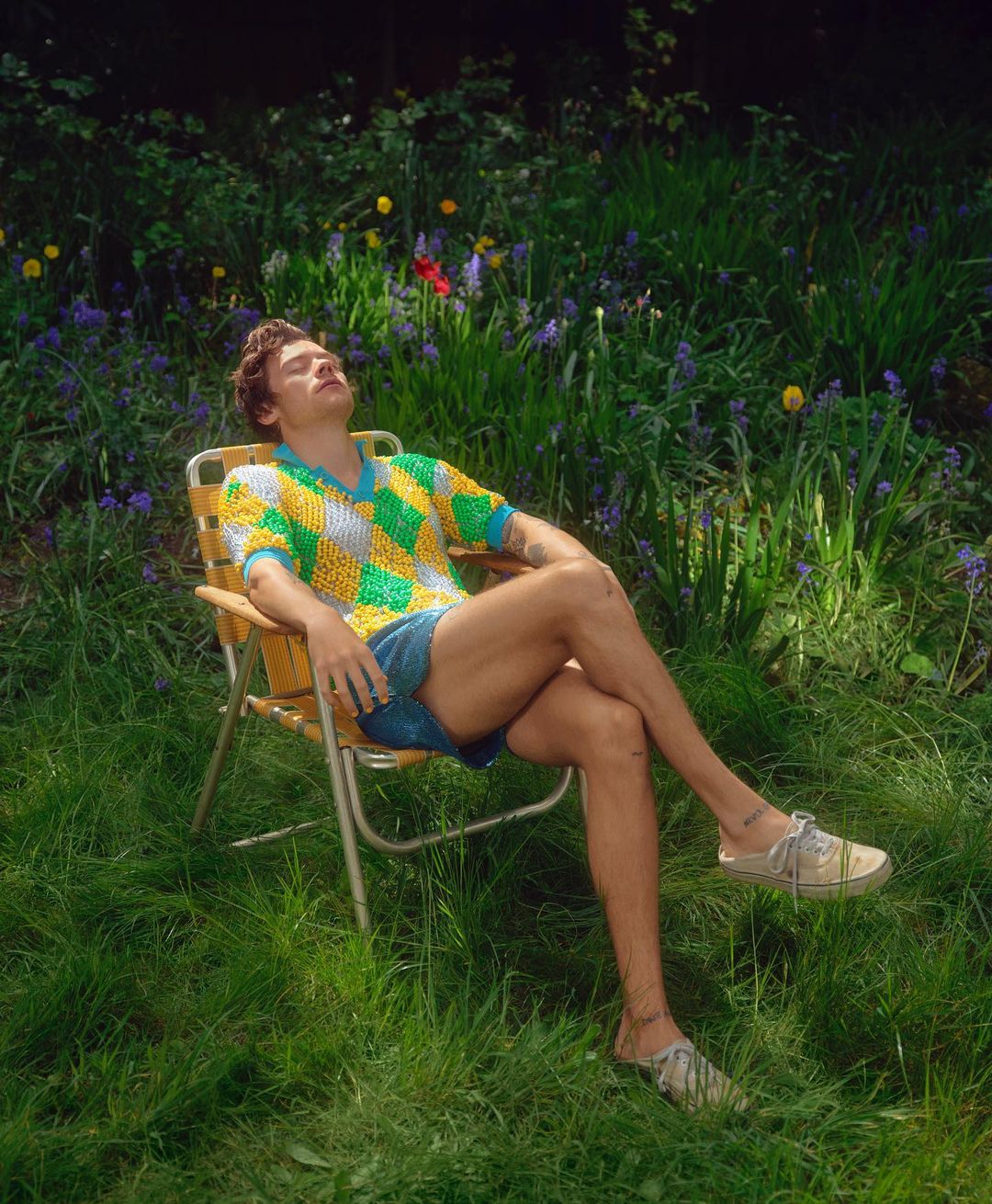 Man relaxes in chair in a grassy field wearing a yellow and green sweater, blue shorts, and beige shoes surrounded by greenery.