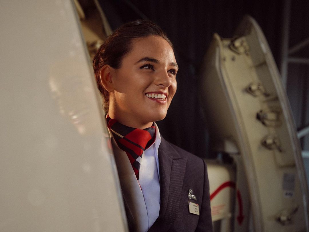 Smiling flight attendant in navy suit with red scarf stands near airplane door in interior shot with natural lighting.