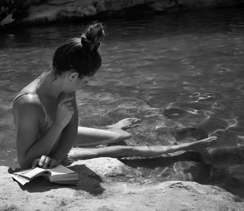 Black and white photo of a young woman in a swimsuit reading a book while sitting in shallow water on a sunny day outdoors.