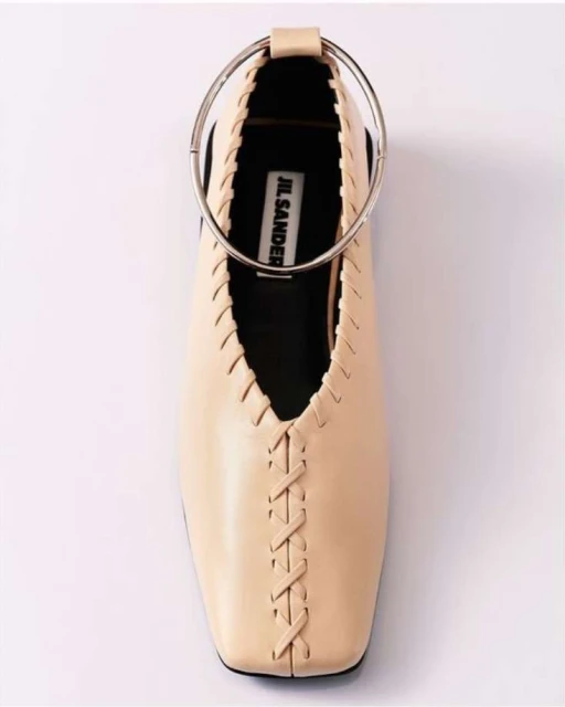 Elevated studio shot focuses on a Jil Sander ballet flat featuring a round ankle strap, intricate detailing, and a soft beige hue.