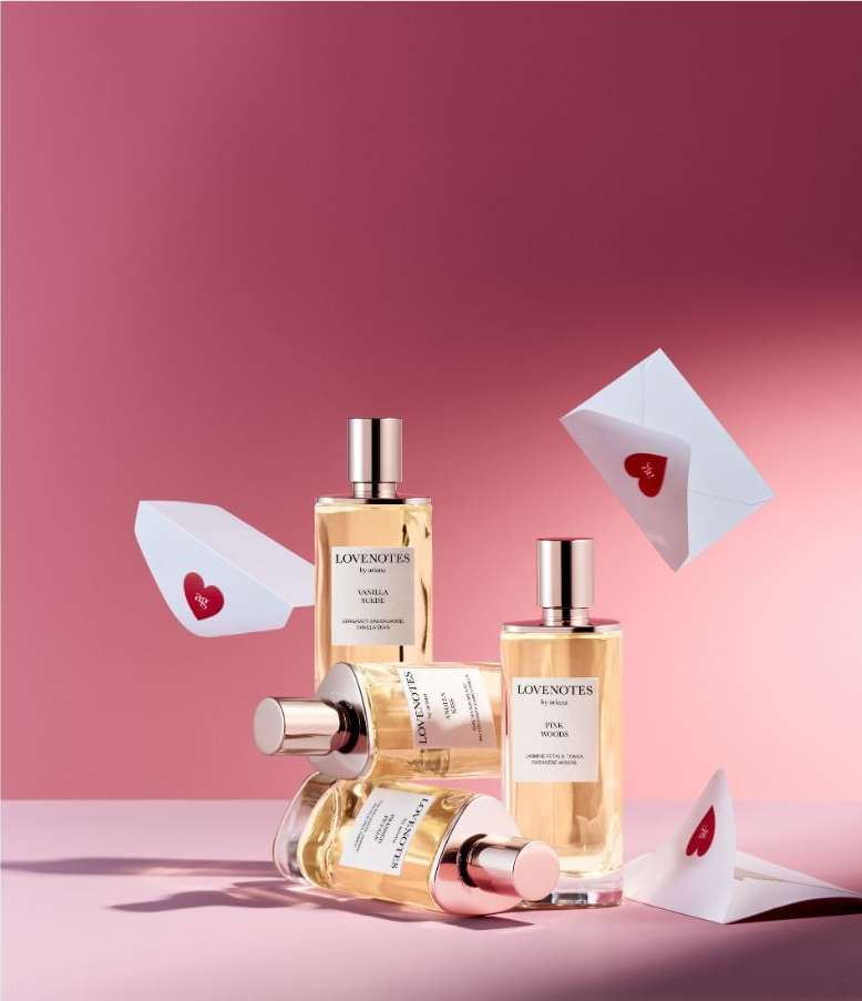 Four perfume bottles arranged against a pink backdrop with two white envelopes sealed with red heart stickers floating nearby.