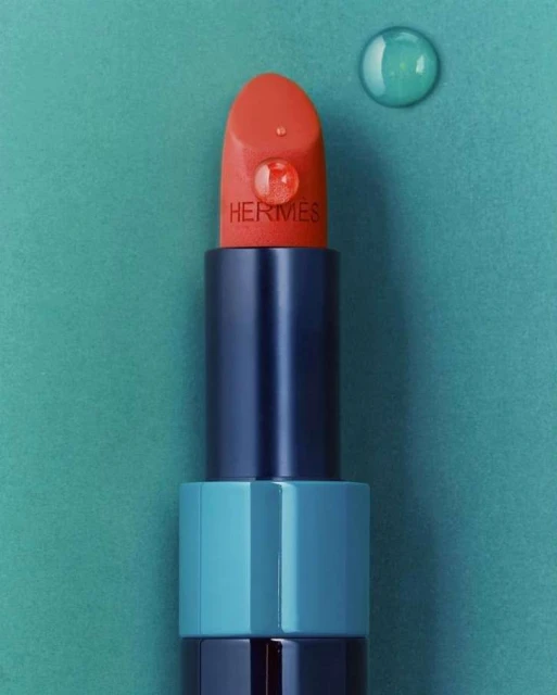 Close-up of an open Hermes lipstick with a water droplet on it, set against a teal background with another water droplet to the side.