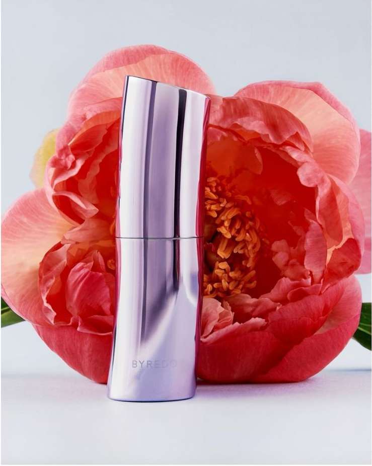 Metallic Byredo perfume bottle standing in front of a lush pink peony flower on a light background, minimalist product shot.