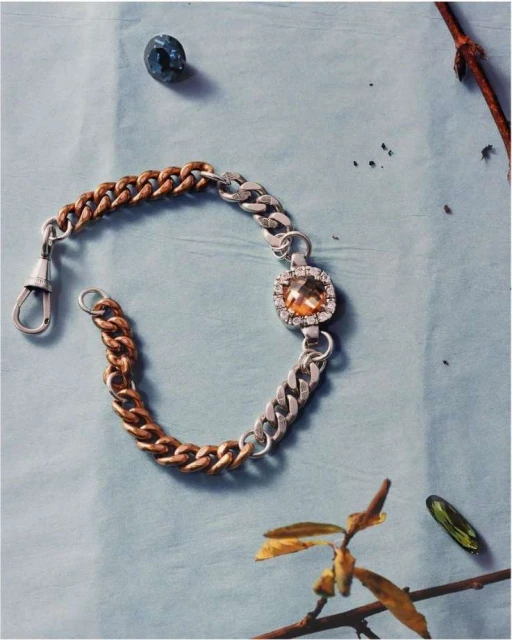 Close-up studio shot of a silver-and-copper chain link bracelet featuring a large, amber-colored square gemstone on a light-blue background.