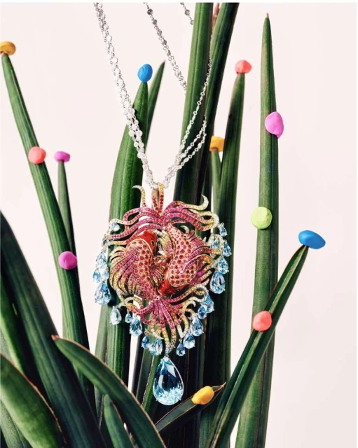 Statement jewelry necklace with pink and blue gems on a silver chain, displayed among green plants with colourful beads.