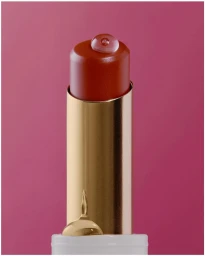 Close up shot of a red lipstick in a gold metallic case with a water droplet on top against a solid pink background.