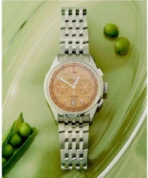A luxury silver chronograph watch with a salmon dial rests on a reflective pale green surface beside whole green peas.