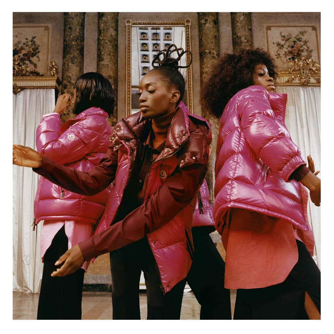 Three African-American women pose indoors wearing pink puffer jackets, black trousers, and long-sleeved shirts in front of a columned wall.
