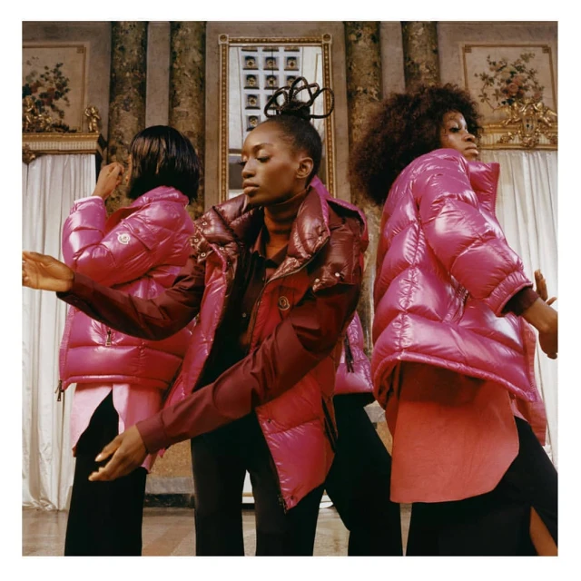 Three African-American women pose indoors wearing pink puffer jackets, black trousers, and long-sleeved shirts in front of a columned wall.
