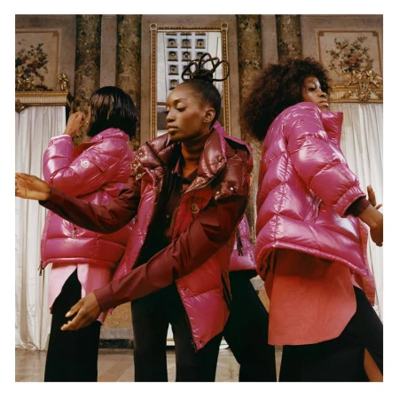 Three African-American women pose indoors wearing pink puffer jackets, black trousers, and long-sleeved shirts in front of a columned wall.