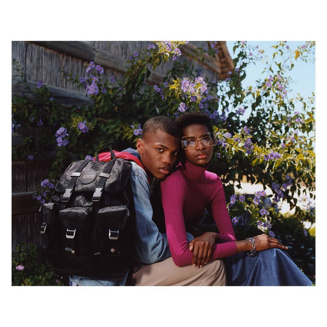Two young adult fashion models pose with a dark Gucci backpack in front of purple flowers and wood paneling outdoors.