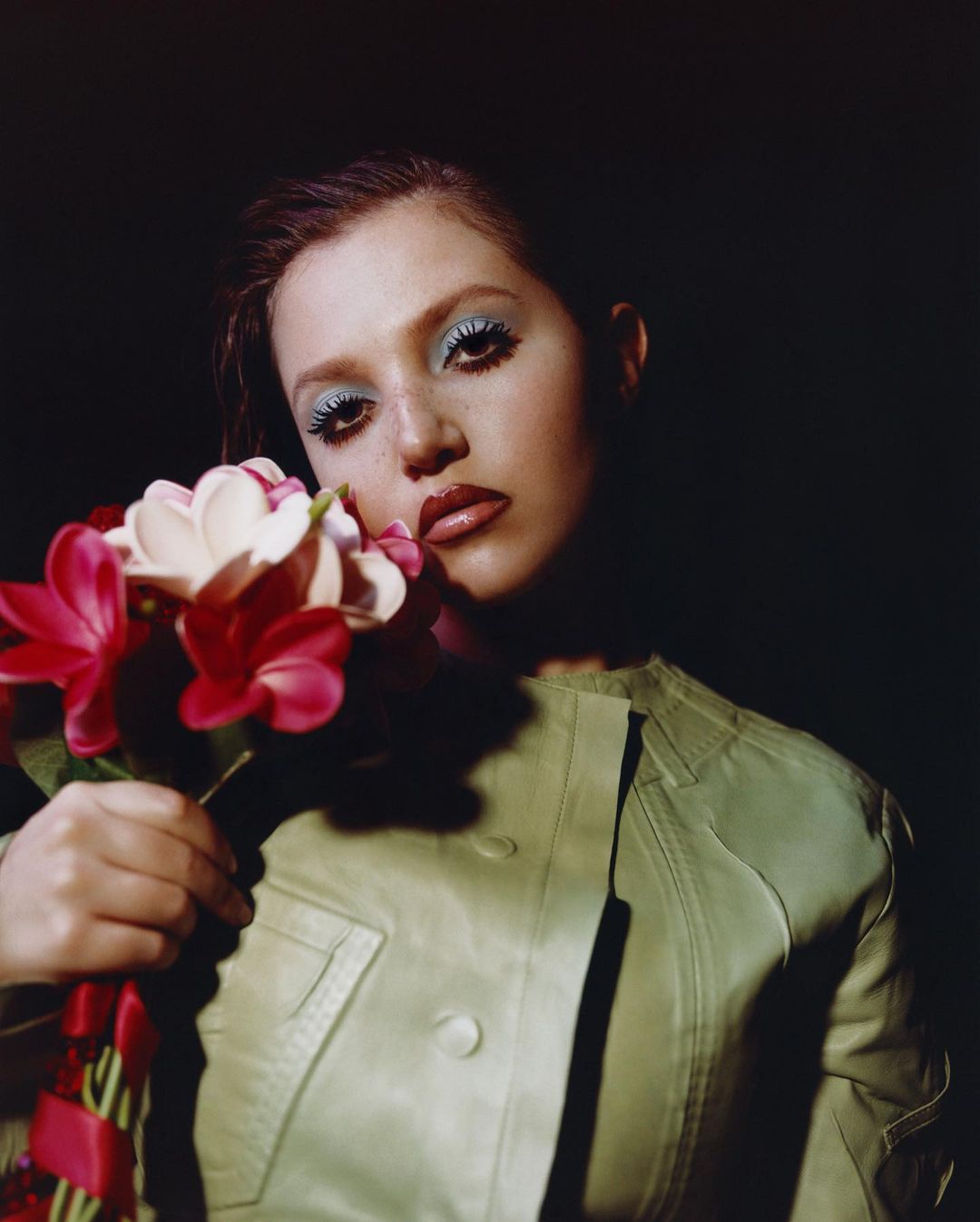 Portrait of a young adult woman holding a bouquet with red and white flowers, wearing a light green jacket against a dark background.