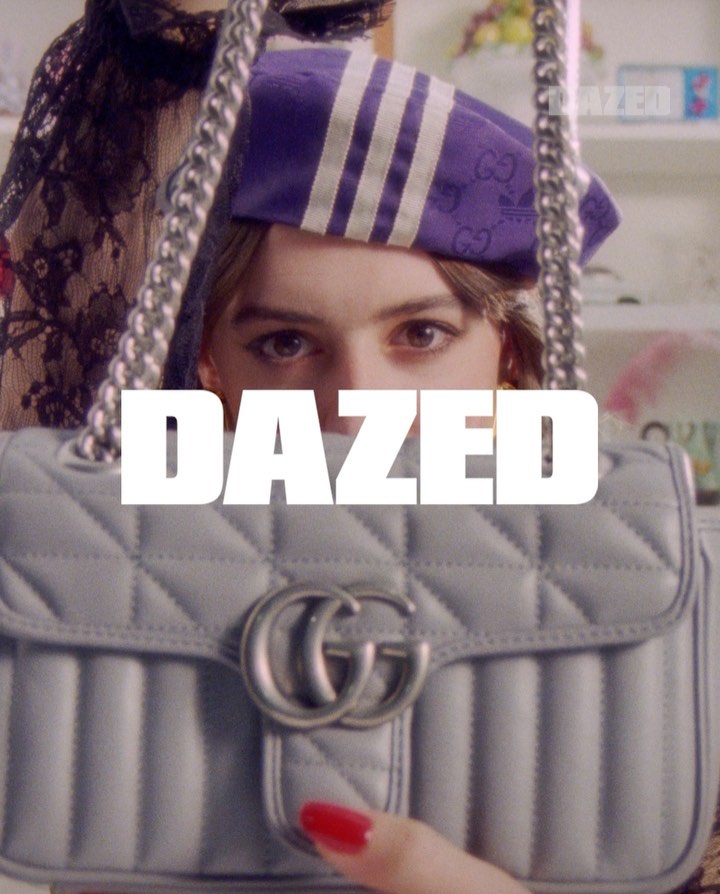 A woman is holding a light blue Gucci purse with the word DAZED in front of her face. She is wearing a purple and white hat.