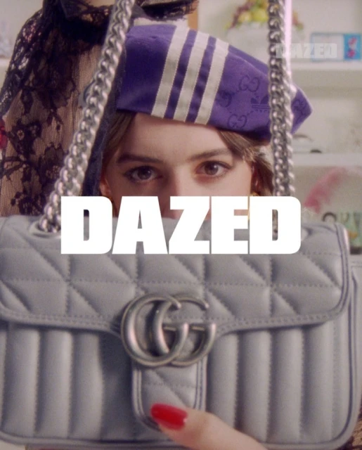 A woman is holding a light blue Gucci purse with the word DAZED in front of her face. She is wearing a purple and white hat.