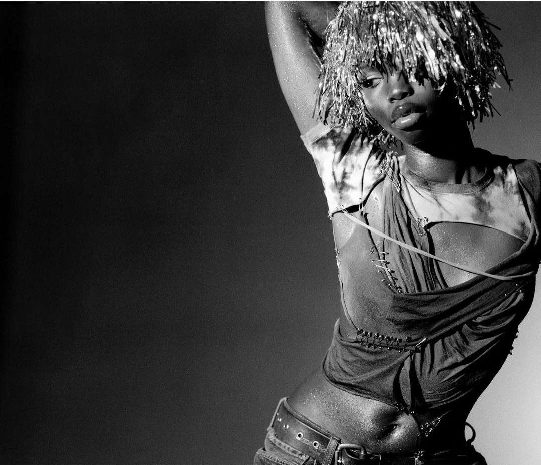 Black woman posing with metallic tinsel wig, wearing distressed tank top and jeans with a belt in a photo studio against a grey background.