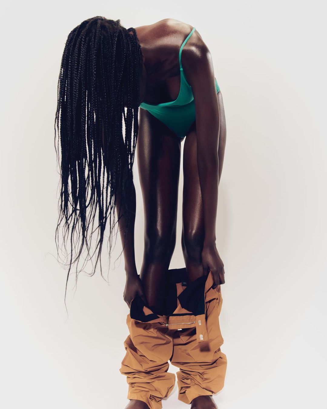 Black woman in a green swimsuit pulling down tan cargo pants in a studio against a white backdrop with long, braided hair.