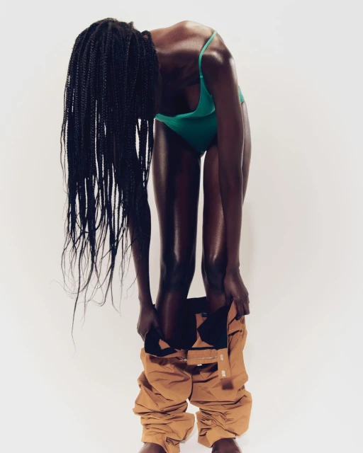 Black woman in a green swimsuit pulling down tan cargo pants in a studio against a white backdrop with long, braided hair.