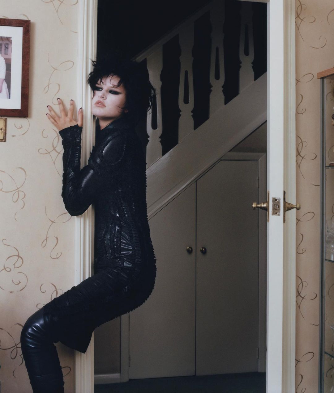 A woman with dark hair and black clothing leans against a door frame, creating a visually striking and slightly surreal portrait.