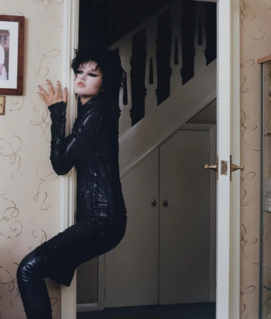 A woman with dark hair and black clothing leans against a door frame, creating a visually striking and slightly surreal portrait.