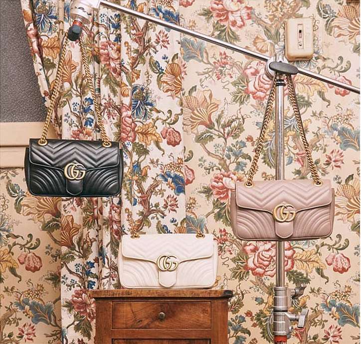Three quilted leather handbags with gold chain straps suspended on metal studio stands against a floral patterned curtain background