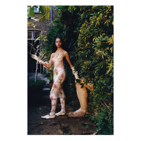 A fashionable woman in a floral gown stands outdoors, complemented by a garden setting and a sculptural leg, blending fashion with nature.
