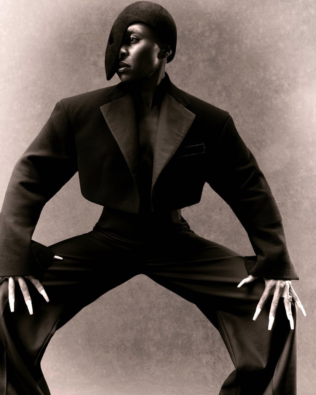 Elegant portrait of a Black woman in a striking black suit and hat, posing in a wide stance against a neutral studio backdrop.