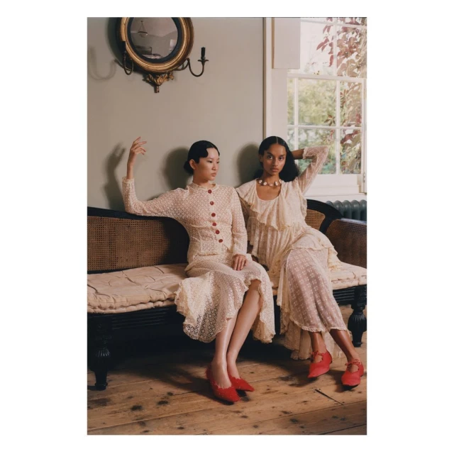 Two elegant women in vintage-inspired dresses pose on a cushioned bench in a room with a window, antique decor, and hardwood floors.