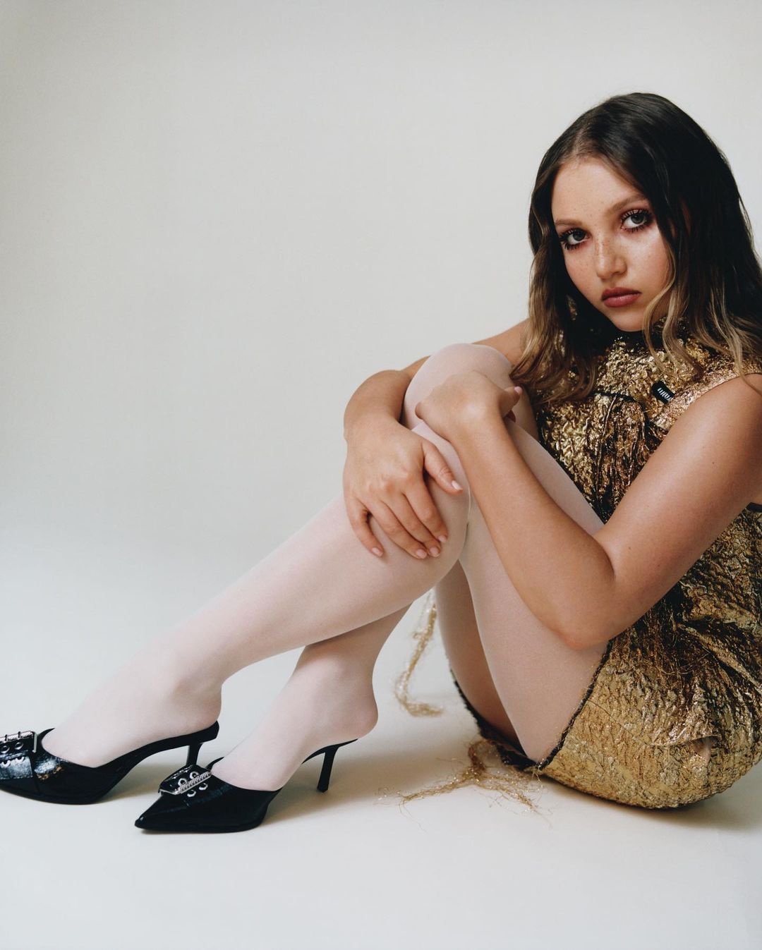 Woman in shimmering gold dress and white tights sitting with legs crossed. Black heeled mules with decorative buckles complete the fashion look.