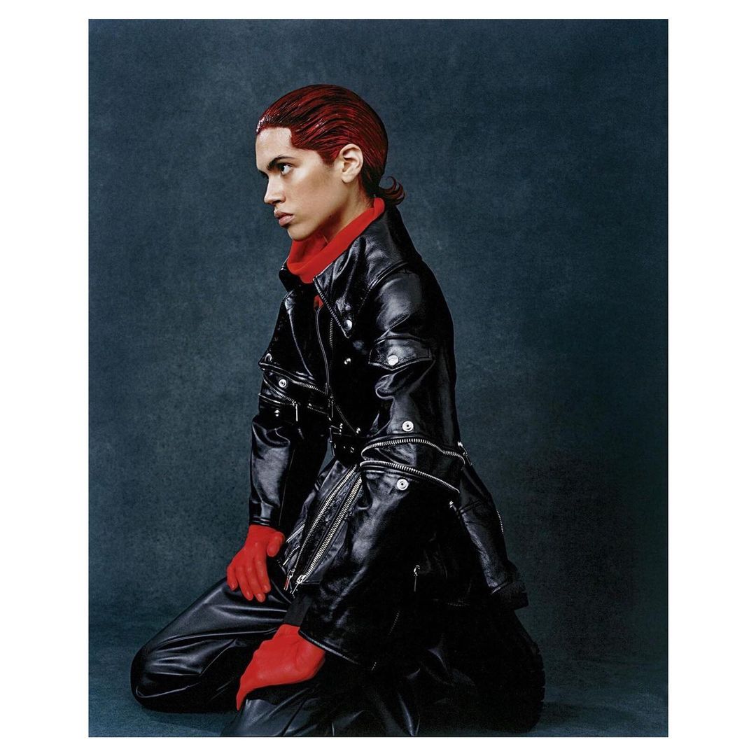 Adult poses on knee in black leather jacket and pants, red turtleneck and gloves against a dark background in a fashion editorial.