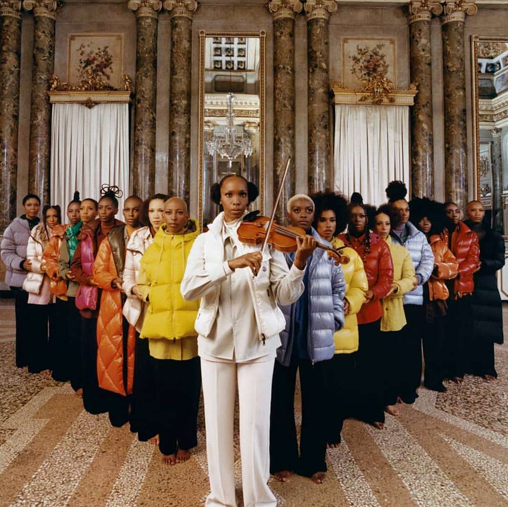 Fashion editorial featuring a group of diverse women in puffer coats standing in a grand, ornate interior, one playing the violin.