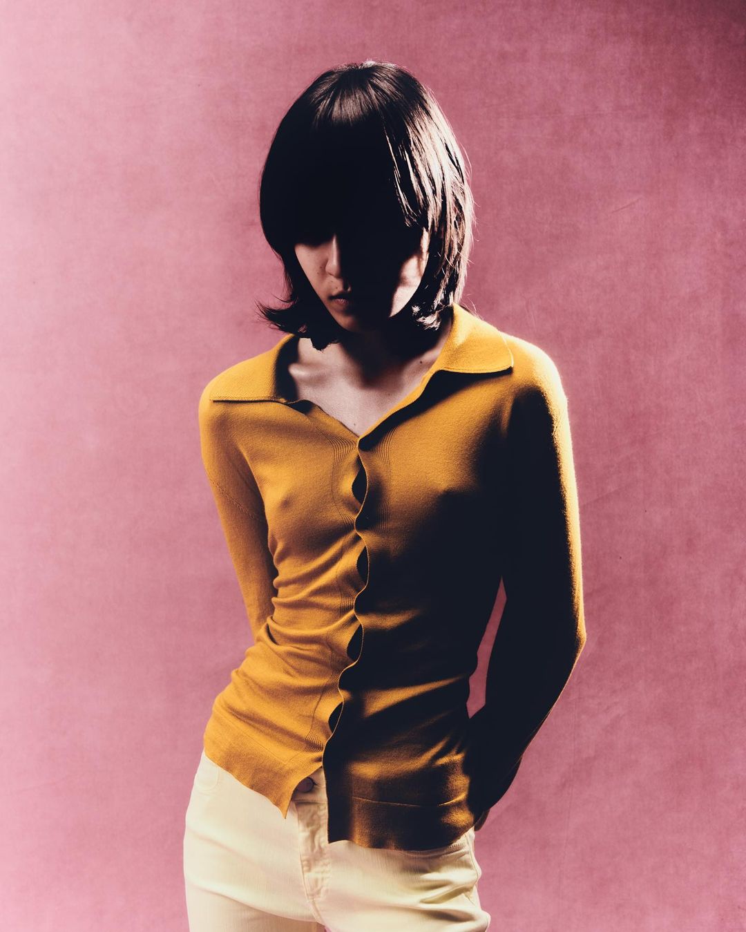 Woman in a yellow collared shirt and white pants stands against a pink backdrop in a studio portrait with strong shadows.
