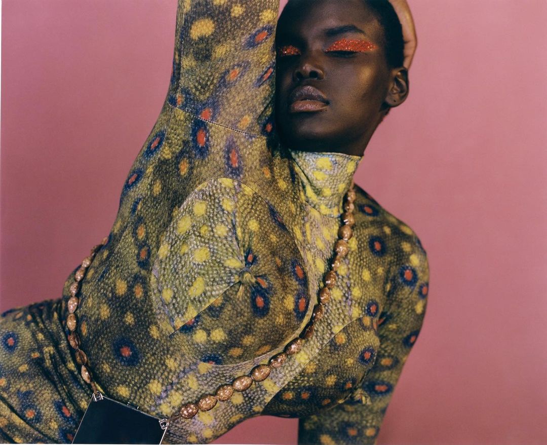 Portrait of a black woman in a yellow-green patterned dress with orange eye makeup, posing against a pink background in a studio setting.