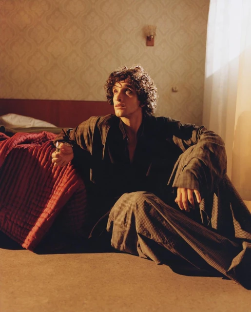 Young man with curly hair sits on the floor in patterned outfit, in a softly lit room with a red bed and patterned wallpaper.