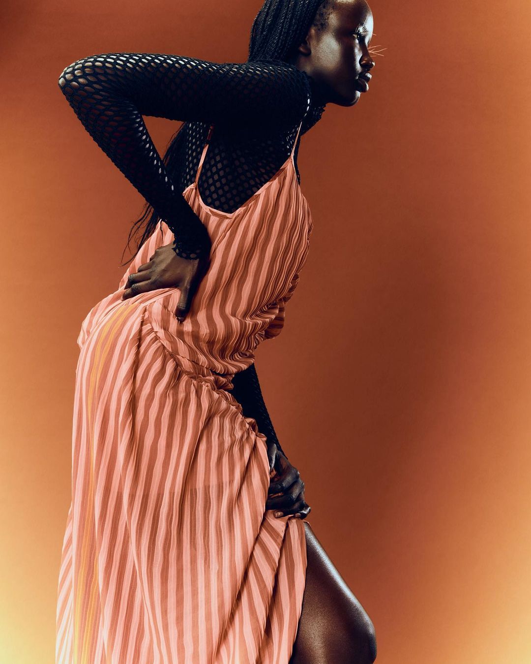Elegant portrait of Black woman in striped dress and mesh top against warm orange backdrop, embodying modern fashion and sophisticated style.