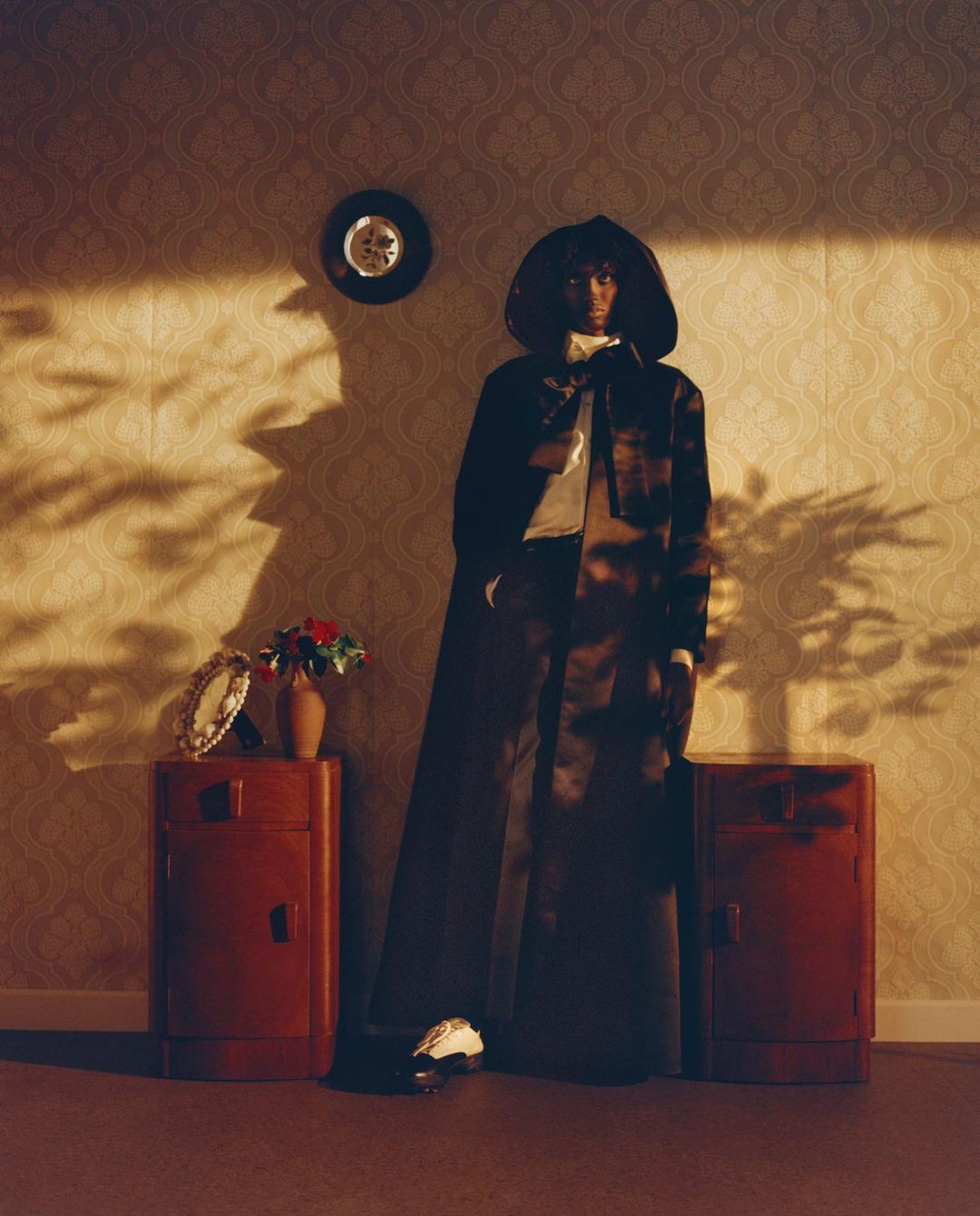 Fashion shot of a woman in a long, dark hooded cloak posing indoors against a patterned wallpaper with side tables and accessories.
