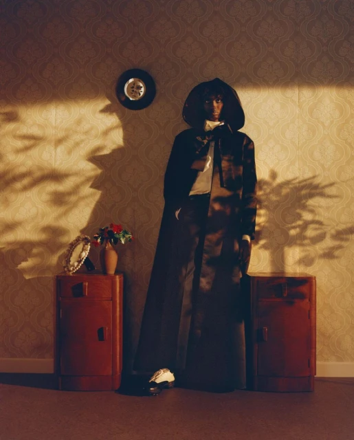 Fashion shot of a woman in a long, dark hooded cloak posing indoors against a patterned wallpaper with side tables and accessories.