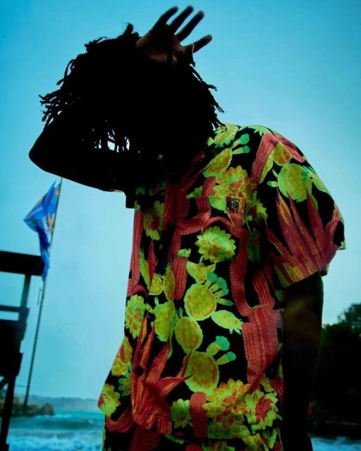 A person in a floral short sleeve shirt stands on a beach under a blue sky, partially obscuring their face with their hand.